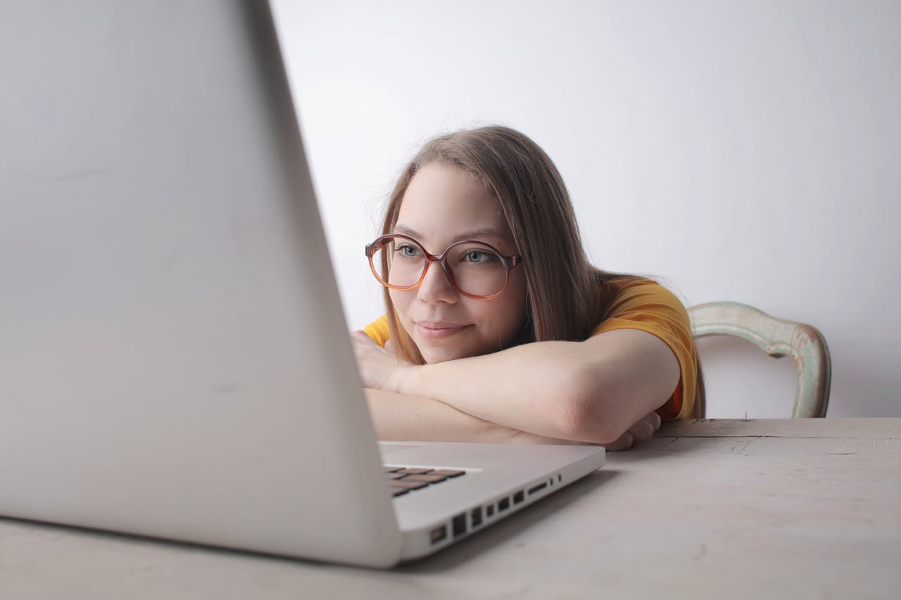A young woman wearing eyeglasses relaxes at home, leaning on a table while using a laptop.