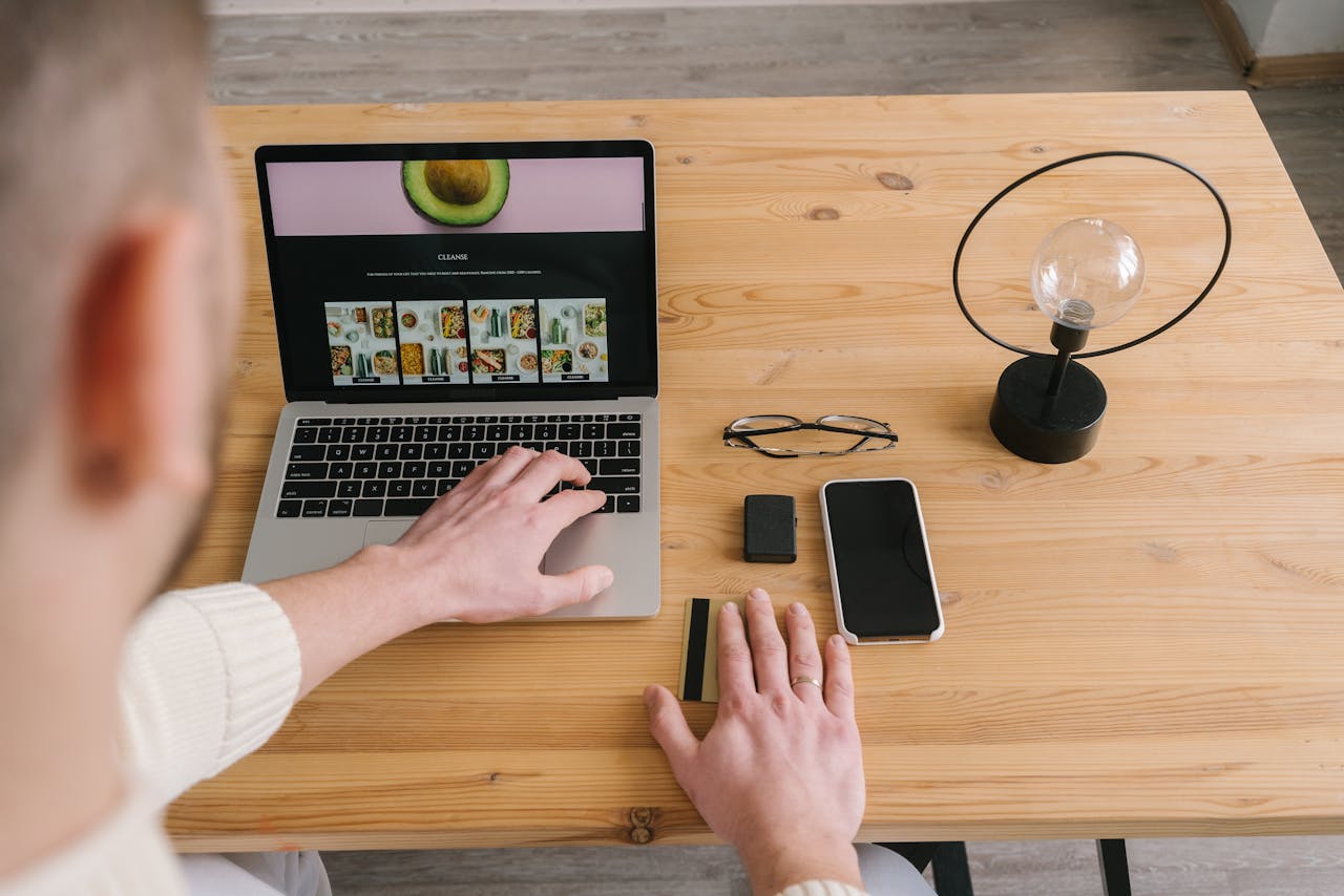 Man using laptop on wooden table with cellphone, bank card, and lamp nearby.
